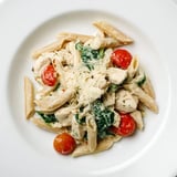 A close-up of Greek Yogurt Chicken Pasta served warm, garnished with fresh basil and a lemon wedge beside a wine glass.  
