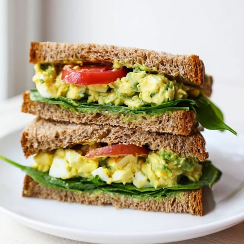 Creamy avocado egg salad sandwich on toasted whole grain bread, layered with fresh spinach and ripe tomato slices for a healthy lunch.